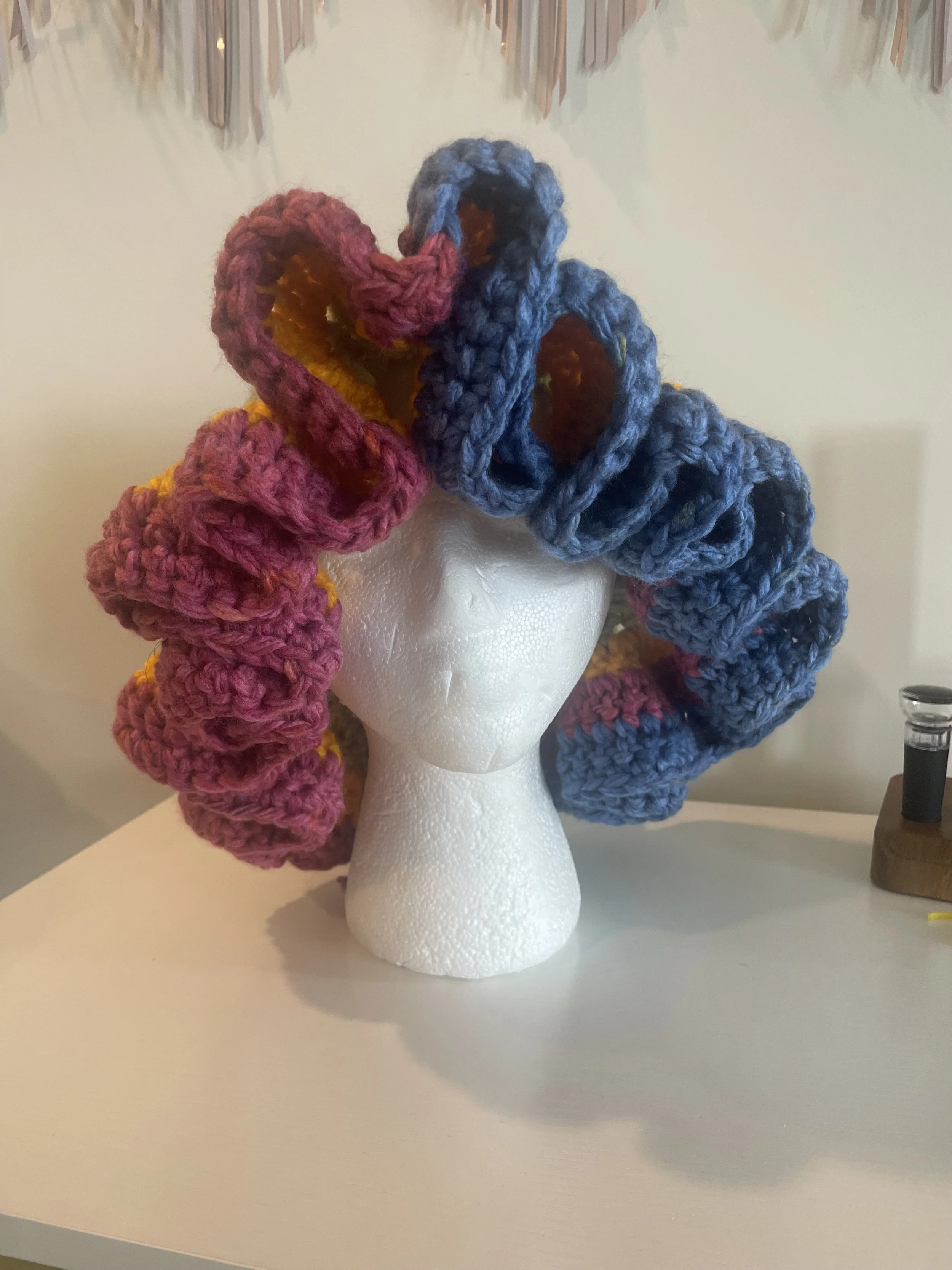 Colorful crocheted loops arranged on a foam mannequin head, creating a wig-like hairstyle, on a white surface.