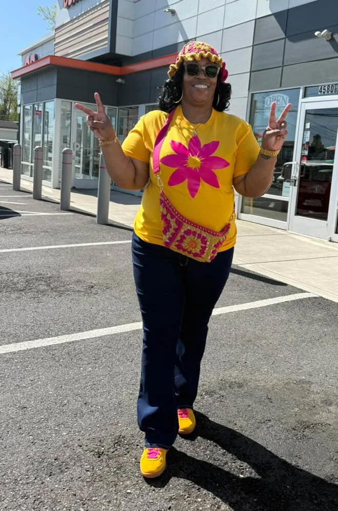 Woman wearing a yellow t-shirt with a large pink flower graphic, yellow and pink shoes, dark jeans, sunglasses, and a yellow crocheted hat with pink accents, standing outside a store in a parking lot, smiling and making peace signs with both hands.