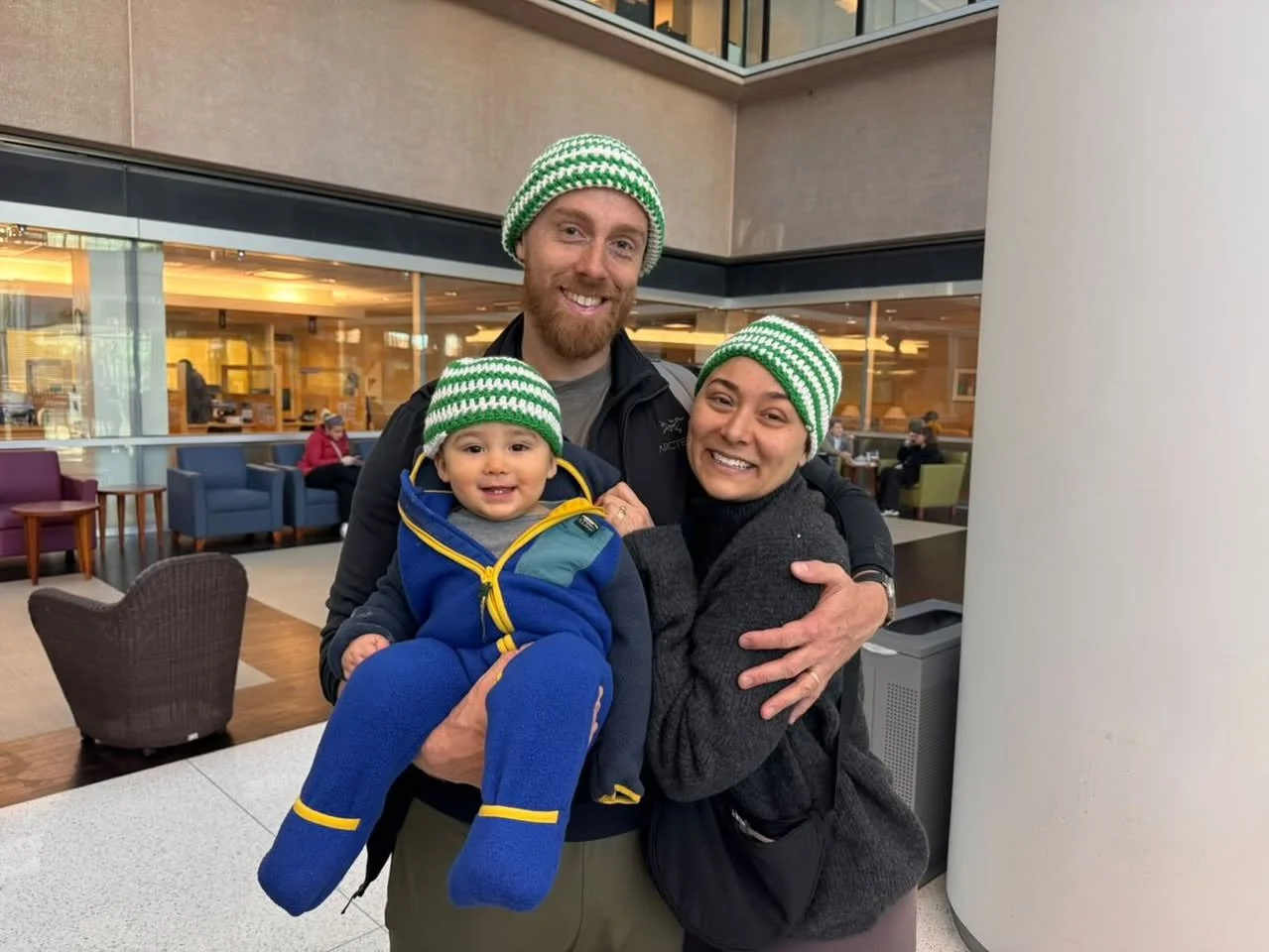 A happy family of three, a man, woman, and a young child, wearing matching green and white striped beanies, posing together in an indoor setting. The man is holding the child, and they are all smiling at the camera.