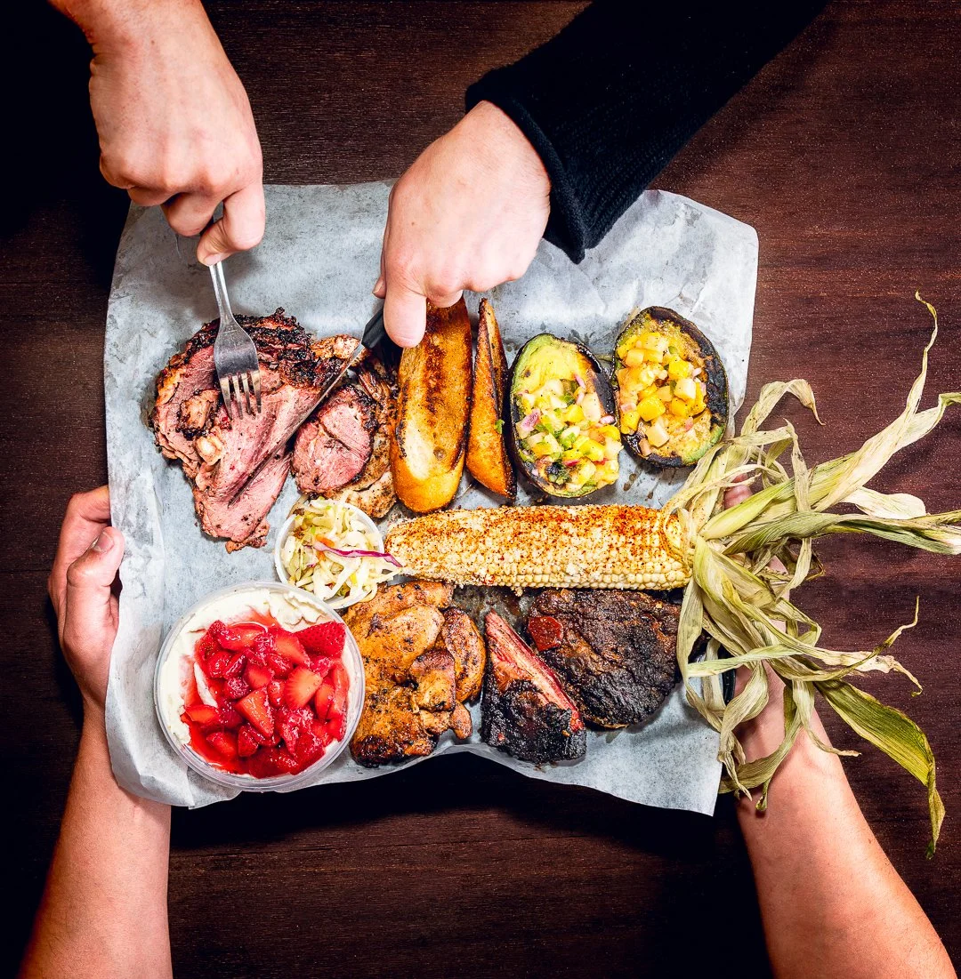 Overhead shot of a sampler platter from Mr. Brown's Brisket Society pop-up in Elk Grove, featuring smoked brisket, grilled corn, charred avocado with mango salsa, sausage, ribs, slaw, and strawberry shortcake. Hands serve the food on parchment paper 