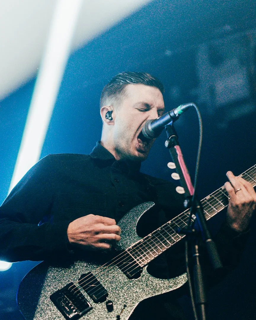 A male musician passionately singing into a microphone while playing an electric guitar on stage, wearing a black shirt.