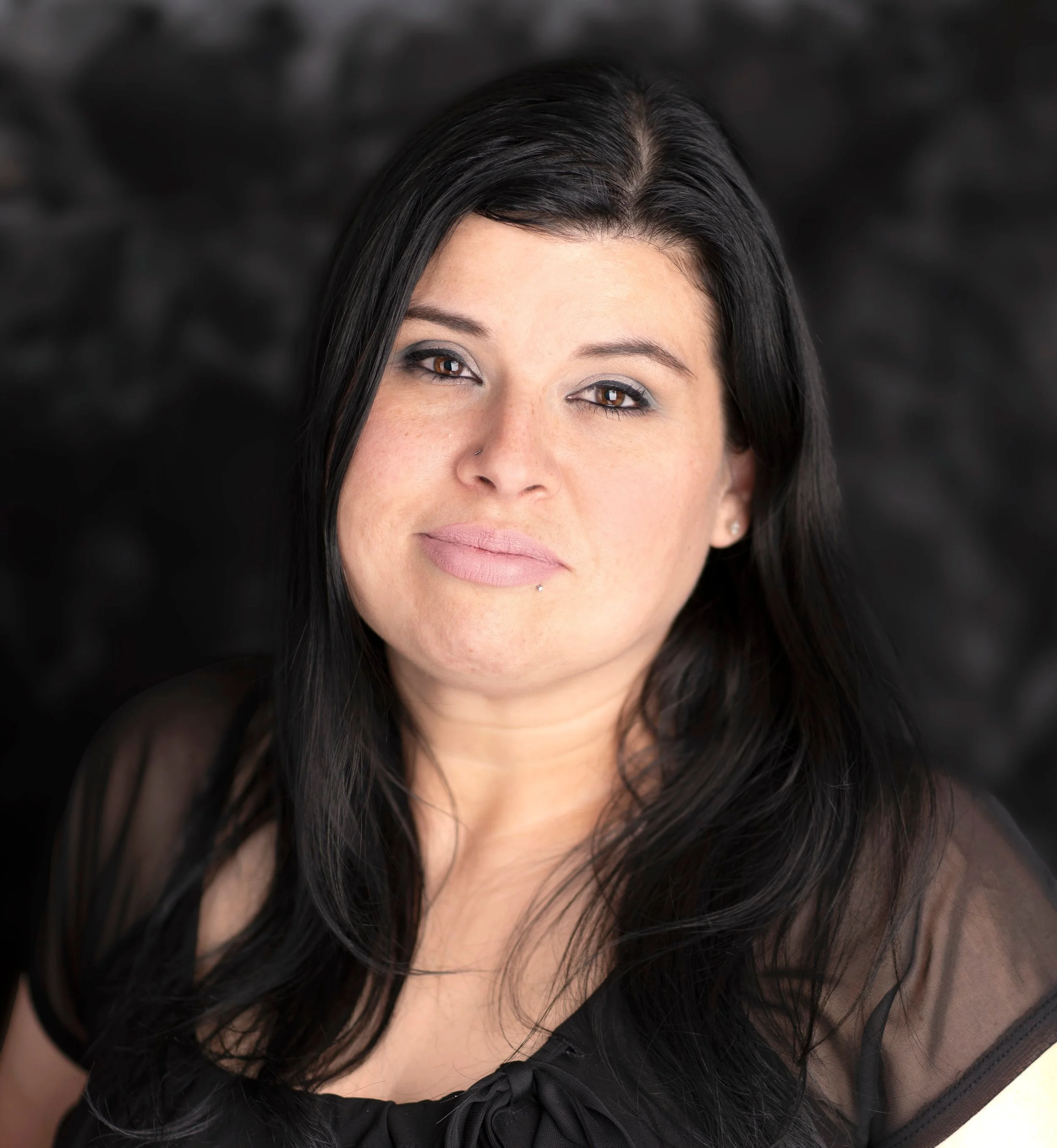 Close-up of a woman with long black hair, light skin, wearing makeup, and a black top with a bow detail, in front of a dark, blurred background.