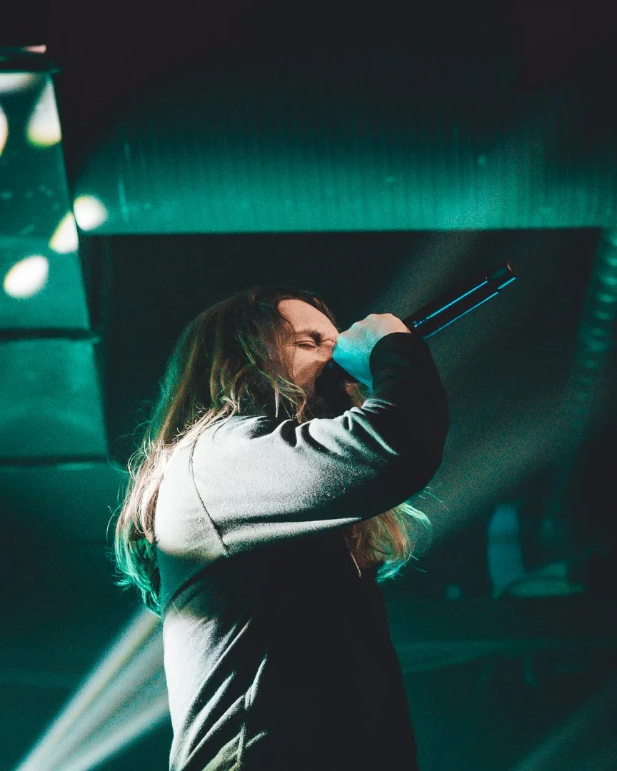 A person with long hair singing into a microphone on stage, eyes closed. They are wearing a dark jacket, with concert lights in the background.