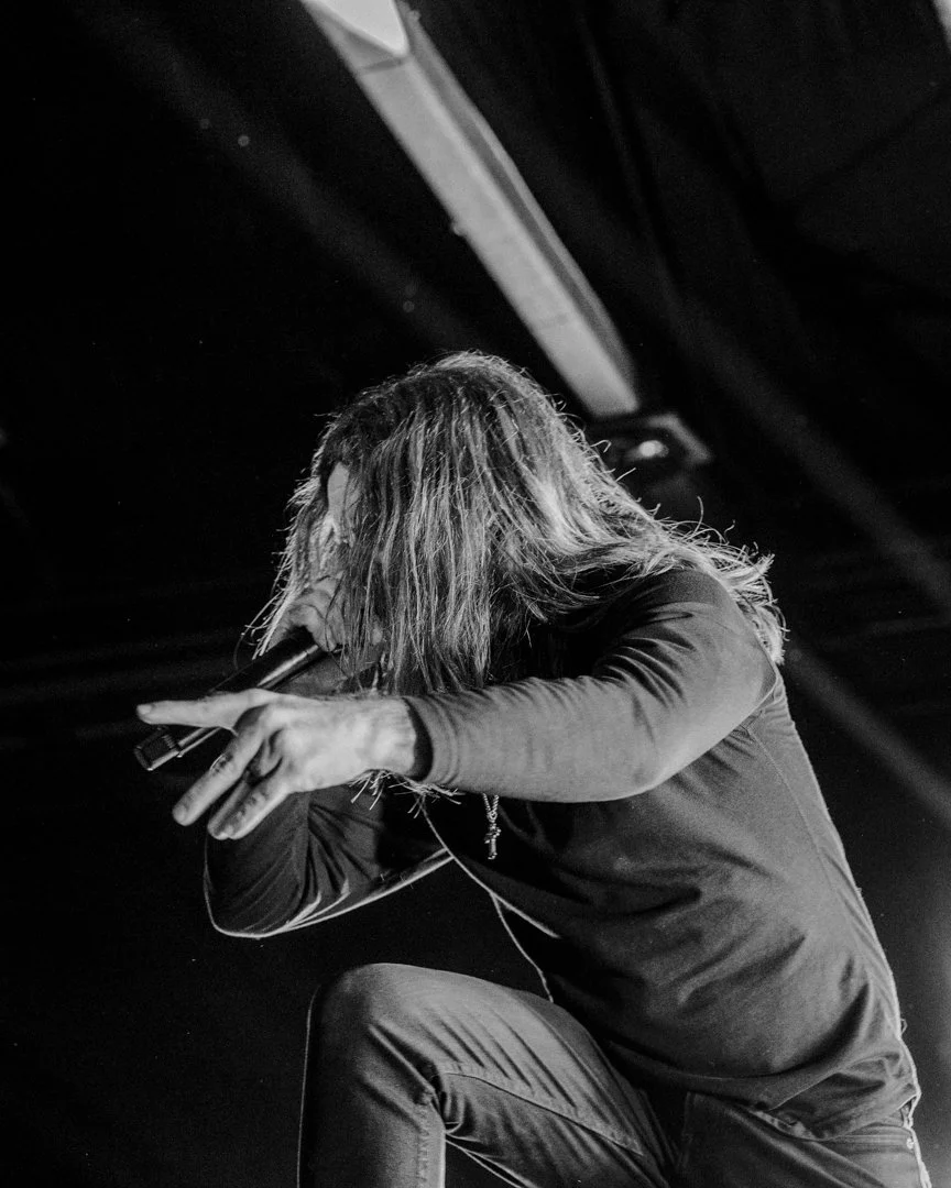 A person with long hair, wearing a dark long-sleeve shirt or jacket, is crouched down on stage, holding a microphone close to their mouth and extending one hand outward, performing in a concert setting.