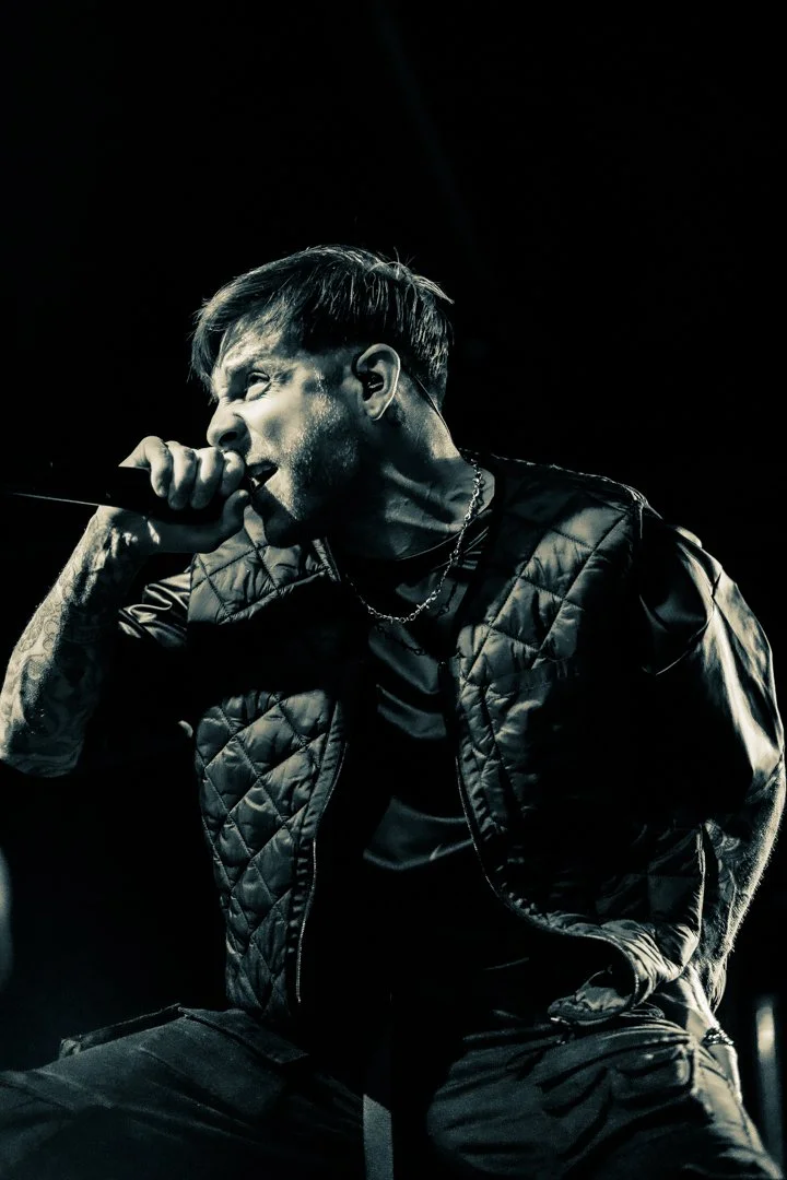 A male singer performing on stage, holding a microphone close to his mouth, wearing a quilted black vest, chain necklace, and tattoos visible on his arm.