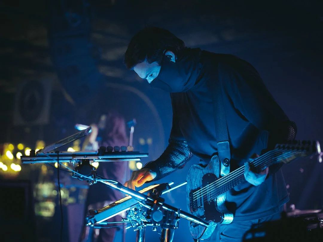 A male musician with dark hair playing an electric guitar on stage, illuminated by blue and yellow stage lights, with equipment and another performer in the background.