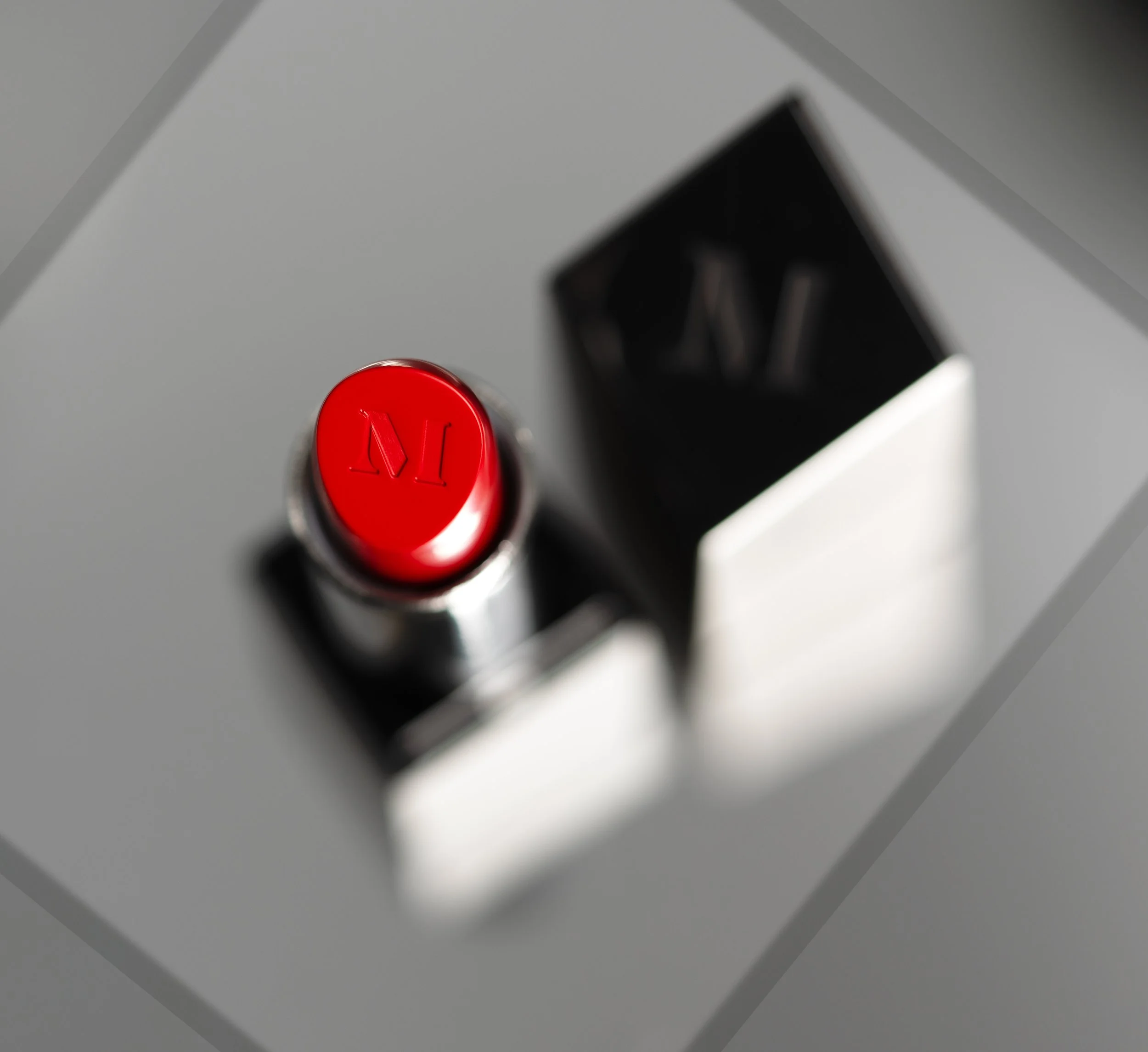 Close-up of a red lipstick with the letter M engraved on its top, placed in a metallic container, with a blurred black and silver box in the background.