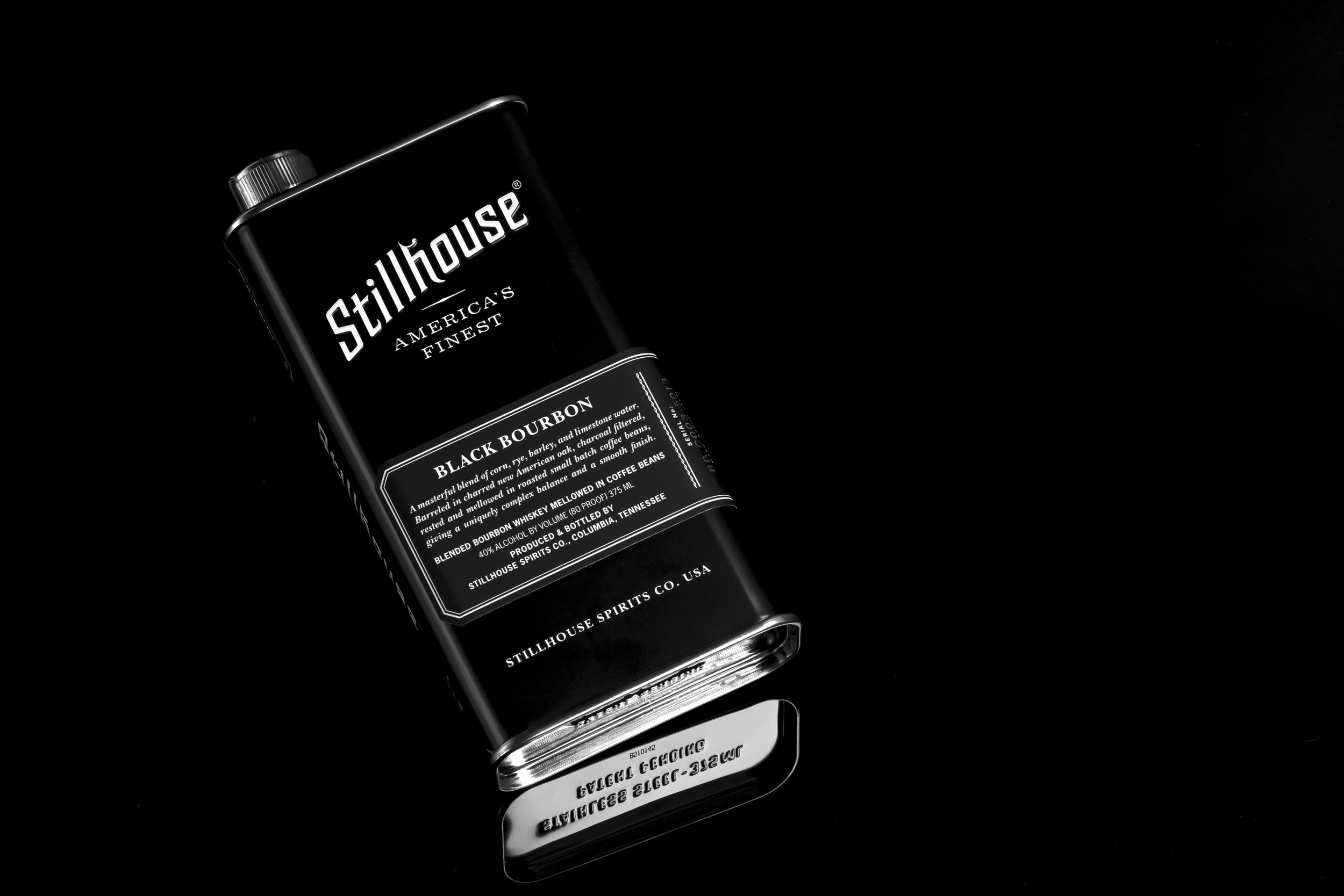 A black and white photograph of a bottle of Stillhouse Black Bourbon whiskey with its label visible, placed on a reflective black surface against a black background.