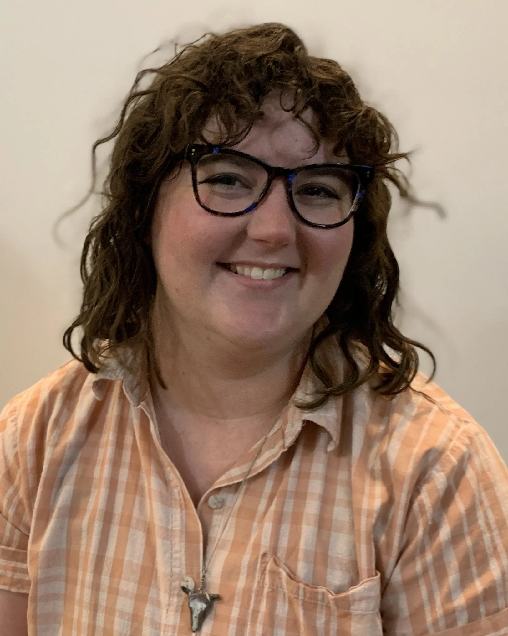 Sarah, smiling. She's in front of a white background, wearing a peachy buttondown, glasses, and a handmade cow skull necklace.
