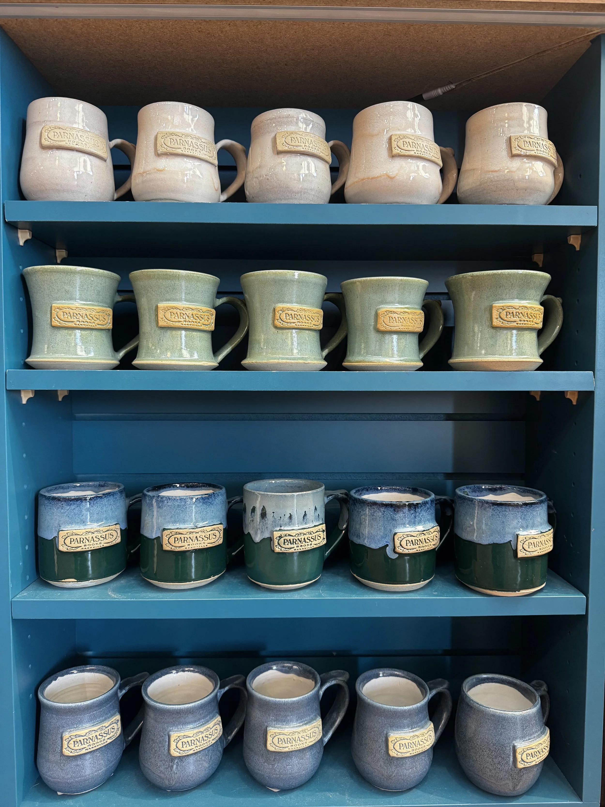 Four rows of mugs in a bookshelf. Branded with the Parnassus Books logo. Made at Old School Farm Pottery, a nonprofit Nashville pottery