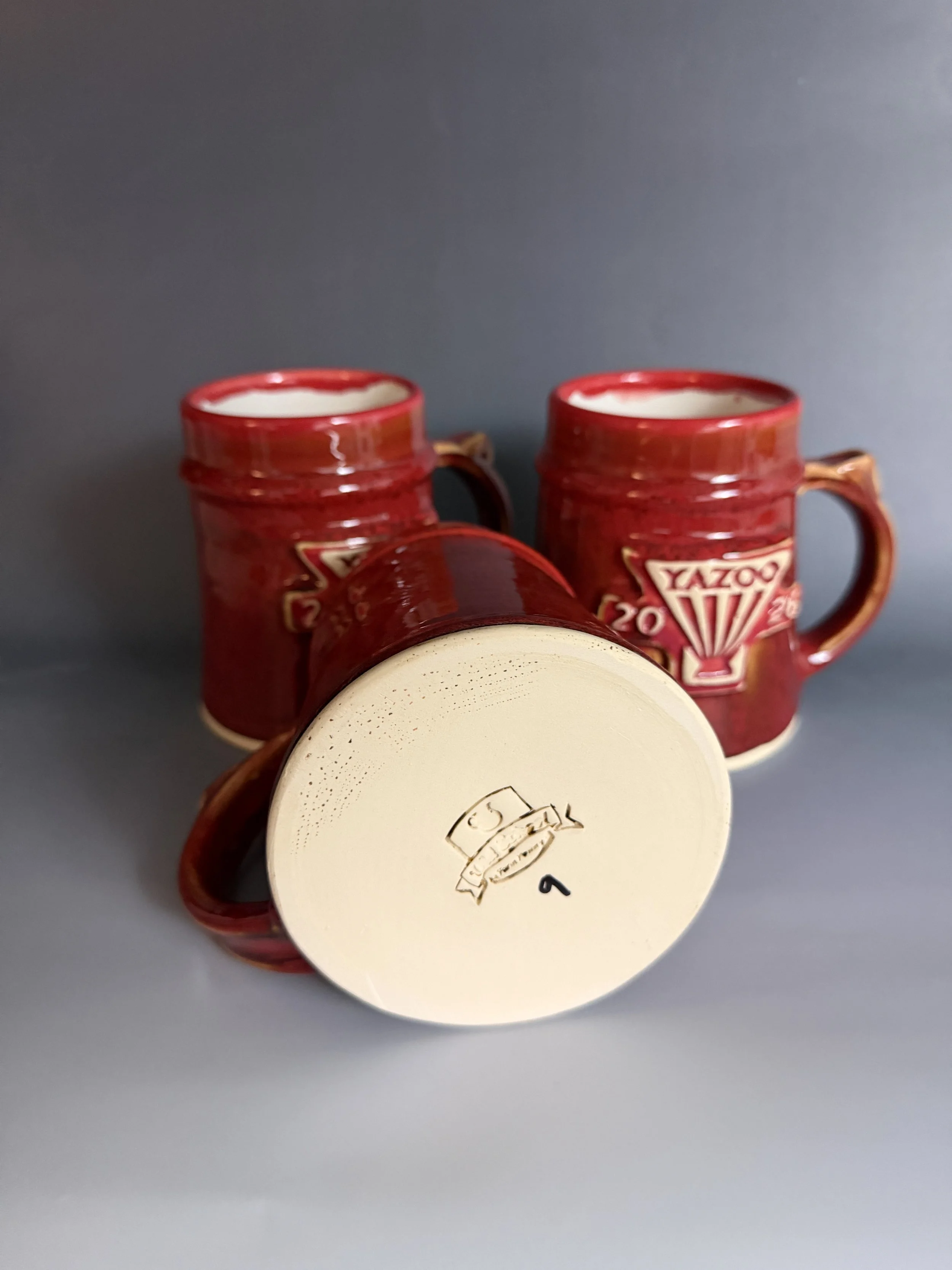 Completed Yazoo 2026 Mug Club Mugs with the bottom of one displayed. Glazed with a custom red and tenmoku glaze. Made at Old School Farm Pottery, a nonprofit Nashville Pottery.