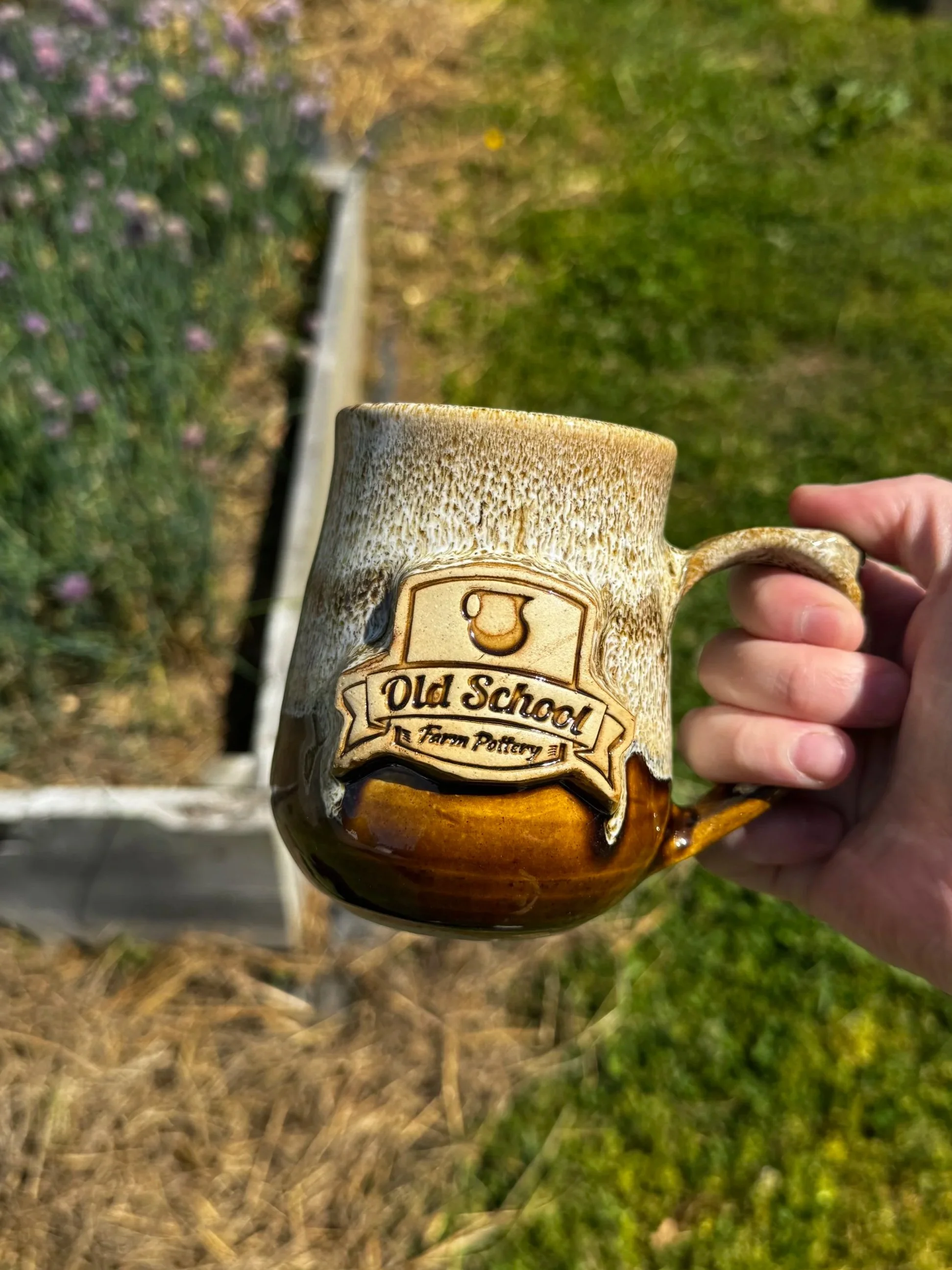 a hand holding a brown and white mug in front of raised garden beds. Log reads "old school farm pottery," which is a nonprofit Nashville pottery