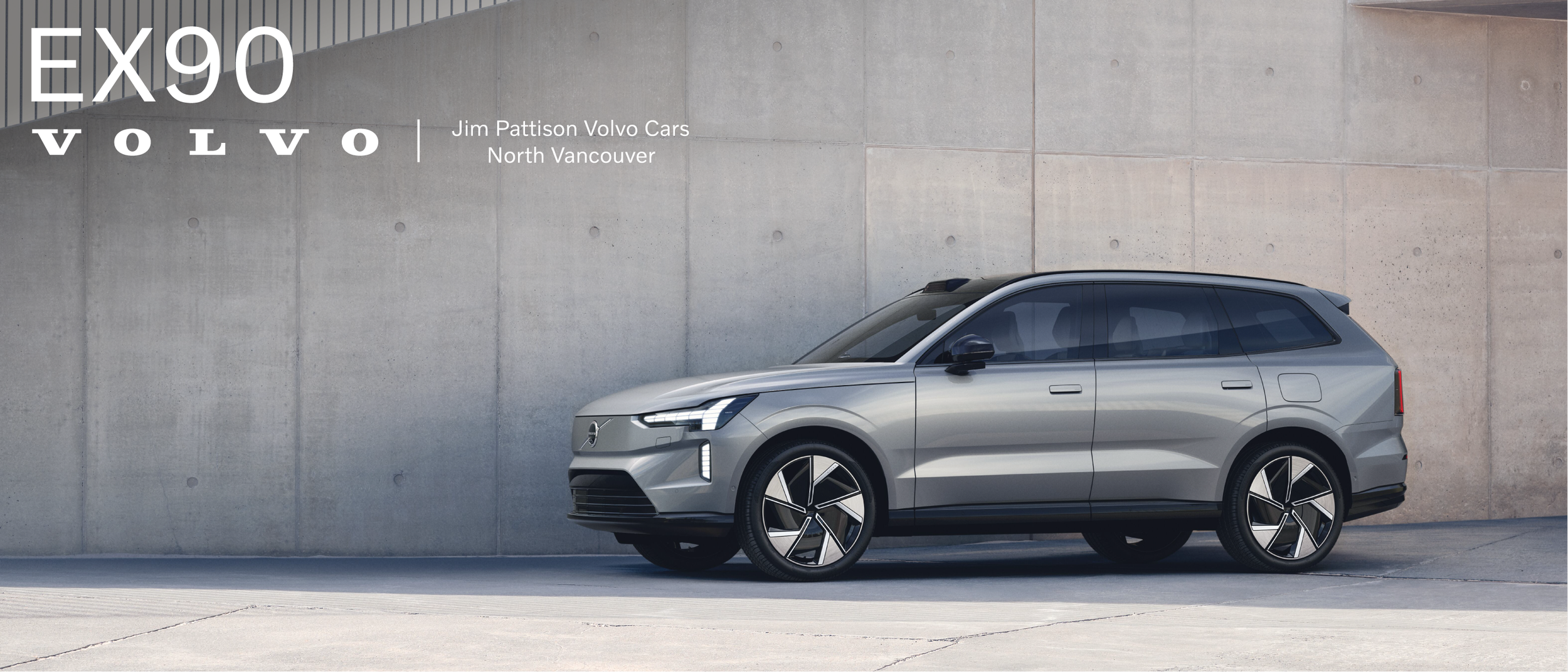A silver Volvo EX90 electric SUV parked against a concrete wall with large text displaying 'EX90' and 'VOLVO', along with dealership information 'Jim Pattison Volvo Cars North Vancouver'.