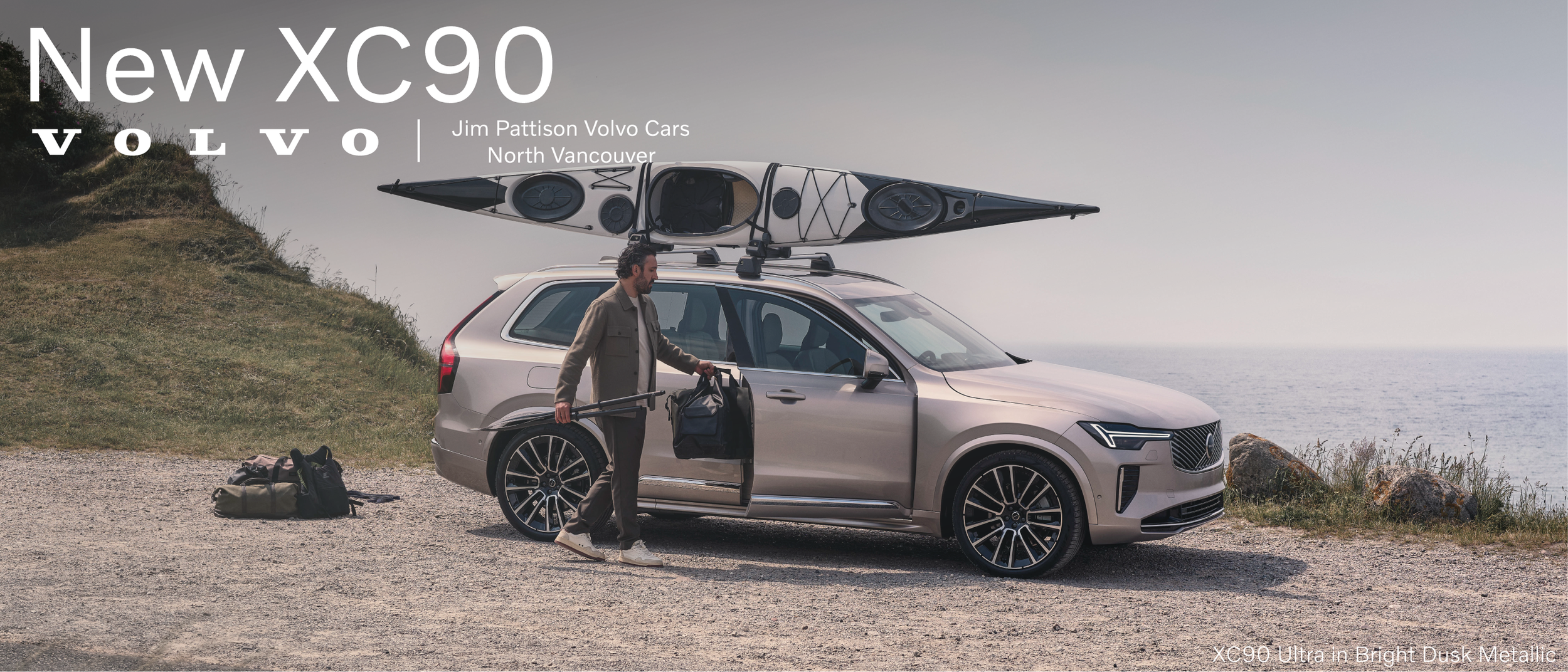 A man preparing for kayaking, standing next to a silver car with a kayak on the roof, by a coast with rocks and grass, with the ocean in the background.