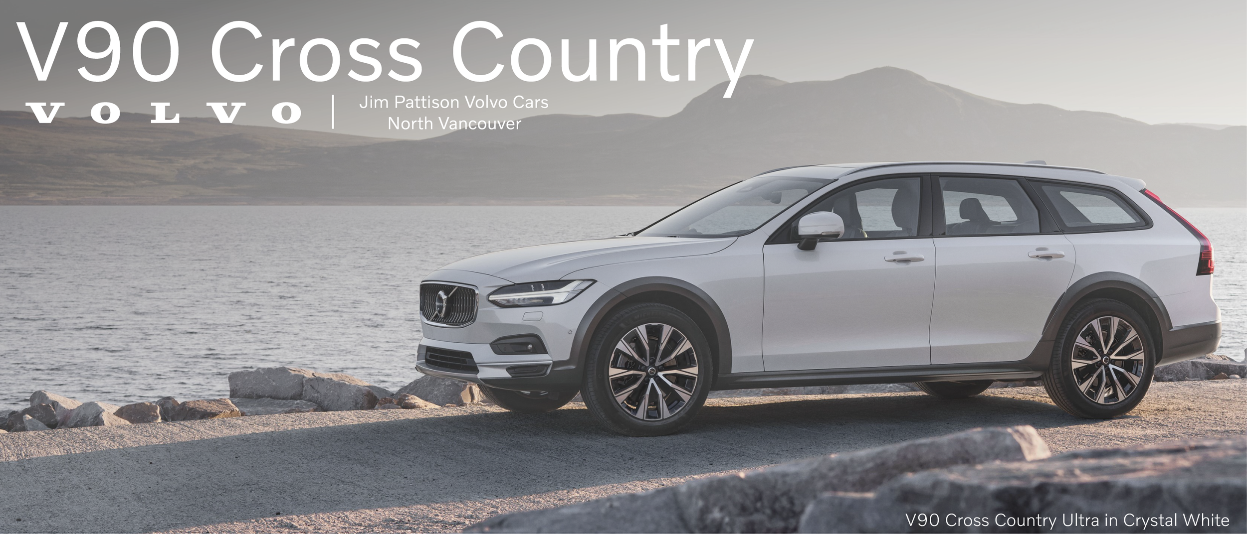 A white Volvo V90 Cross Country station wagon parked by a lake with mountains in the background, promoting the V90 Cross Country model.