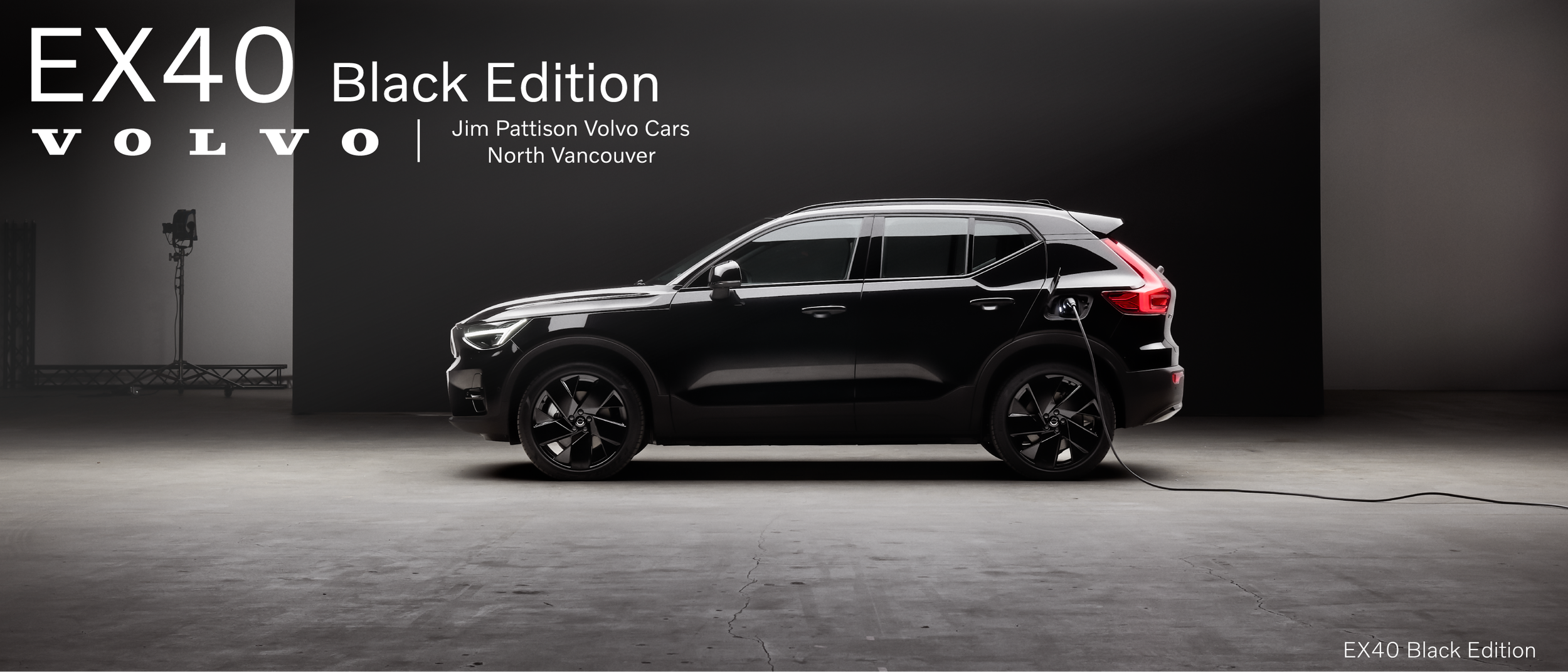 A black electric Volvo SUV connected to a charging cable in a studio setting with a black background and text promoting the EX40 Black Edition from Jim Pattison Volvo Cars North Vancouver.