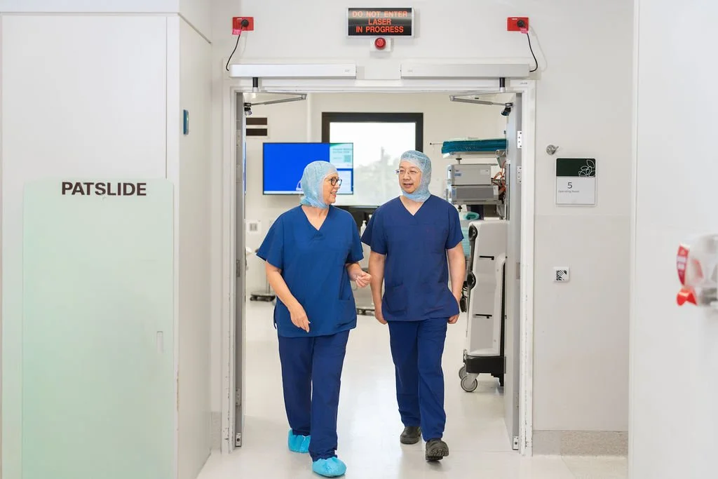 A scrub nurse in navy scrubs and hair covering smiles as she pushes a large Philips X-ray Image Intensifier through the entrance to an operating theatre corridor.