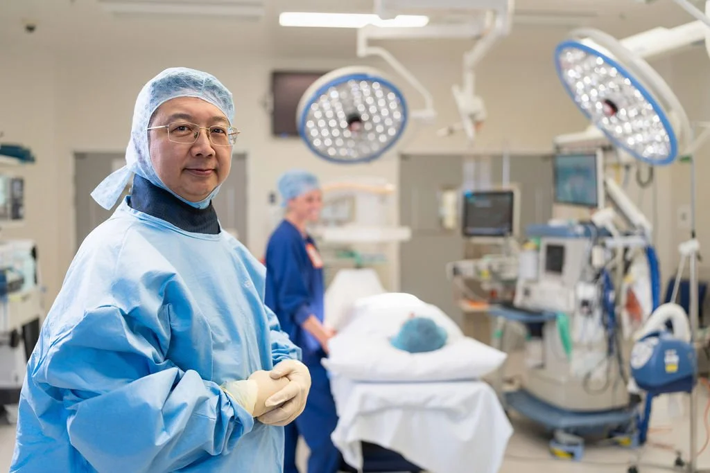 A male orthopaedic surgeon in surgical gown, gloves, and hair covering stands facing camera with a calm, direct expression inside a fully equipped operating theatre.