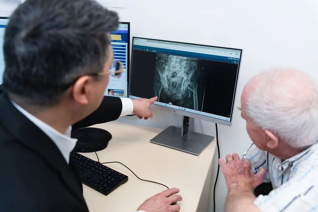 A male orthopaedic surgeon in a dark suit points at a pelvis X-ray on a monitor screen while an elderly male patient looks on, seated beside a desk in a consultation room.