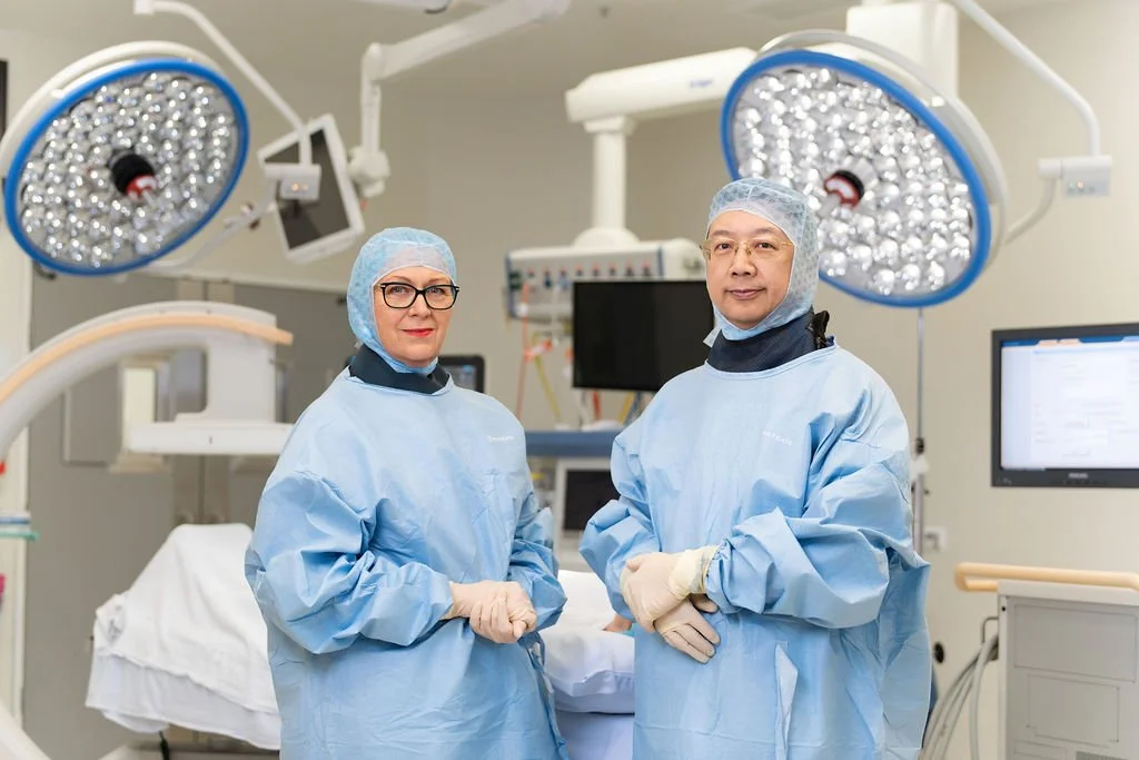 A female and male orthopaedic surgeon stand side by side in surgical gowns and hair coverings inside an operating theatre, looking calmly at camera.