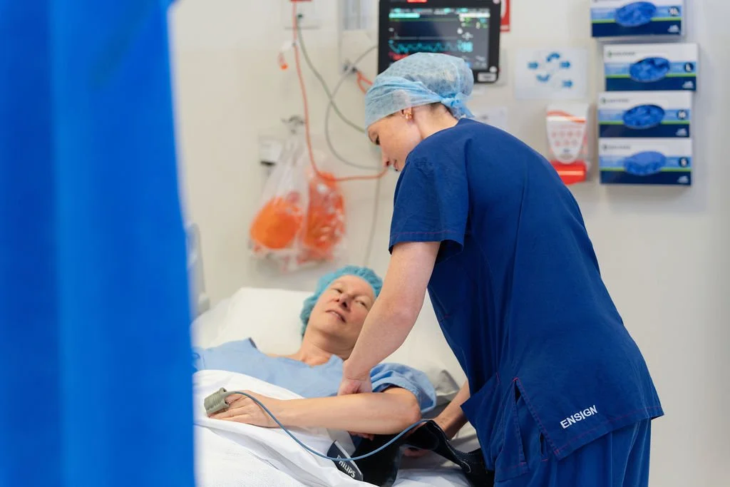 A nurse in navy scrubs and hair covering attaches a pulse oximeter to a patient's finger in a hospital bed, with monitoring equipment and supply drawers visible on the wall behind.
