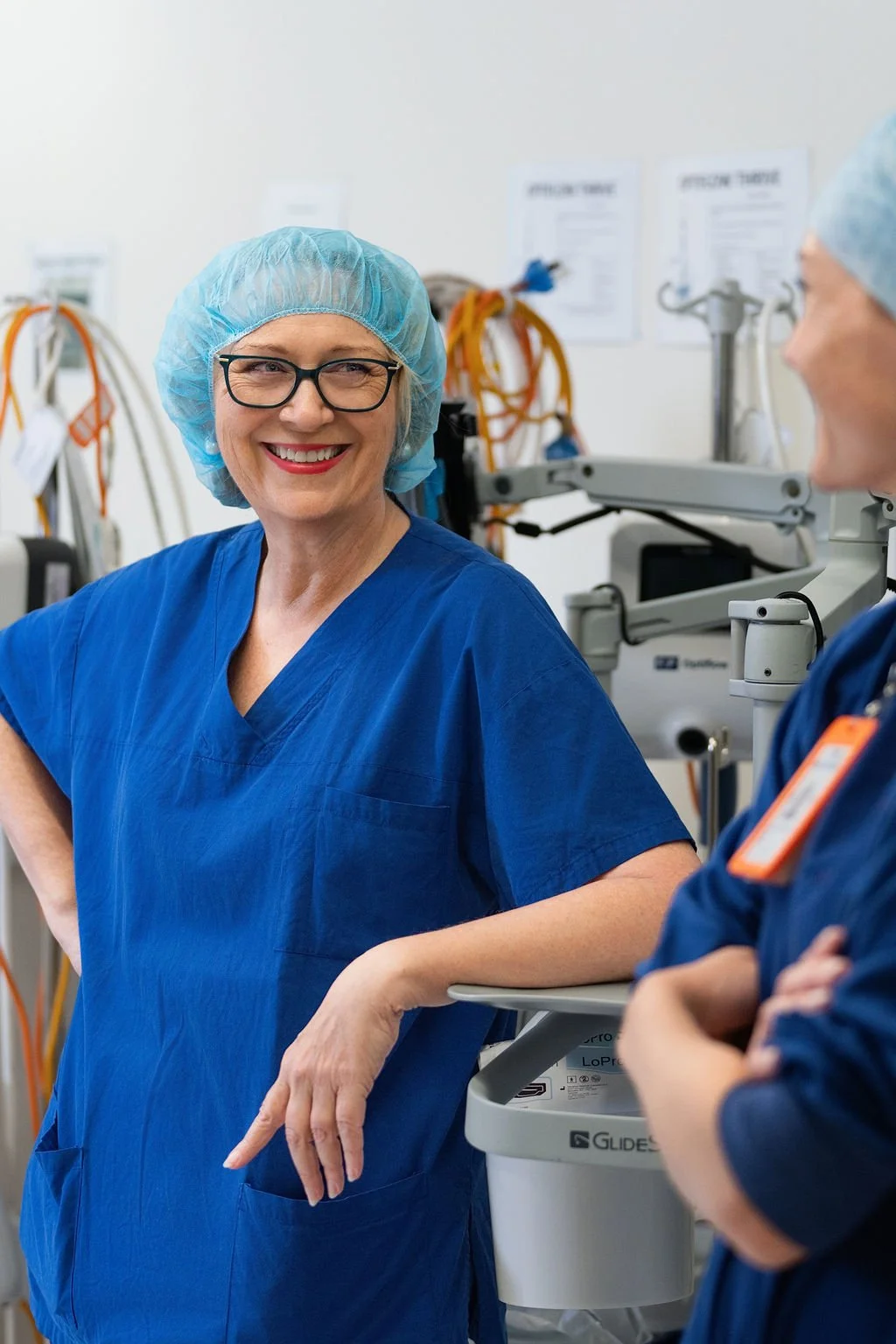 A female orthopaedic surgeon in blue scrubs smiles warmly, leaning against a surgical equipment trolley inside an operating suite, with a colleague partially visible beside her.