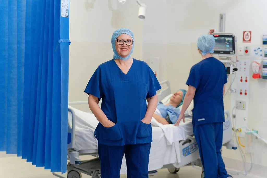 A female nurse in blue scrubs and surgical cap stands with hands in pockets beside a patient bay, smiling calmly at camera, with a nurse and patient visible in the background.