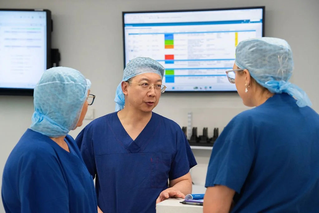 Three surgical team members in navy scrubs and hair coverings stand in a hospital anteroom in discussion, with a colour-coded operating list displayed on the monitor behind them.