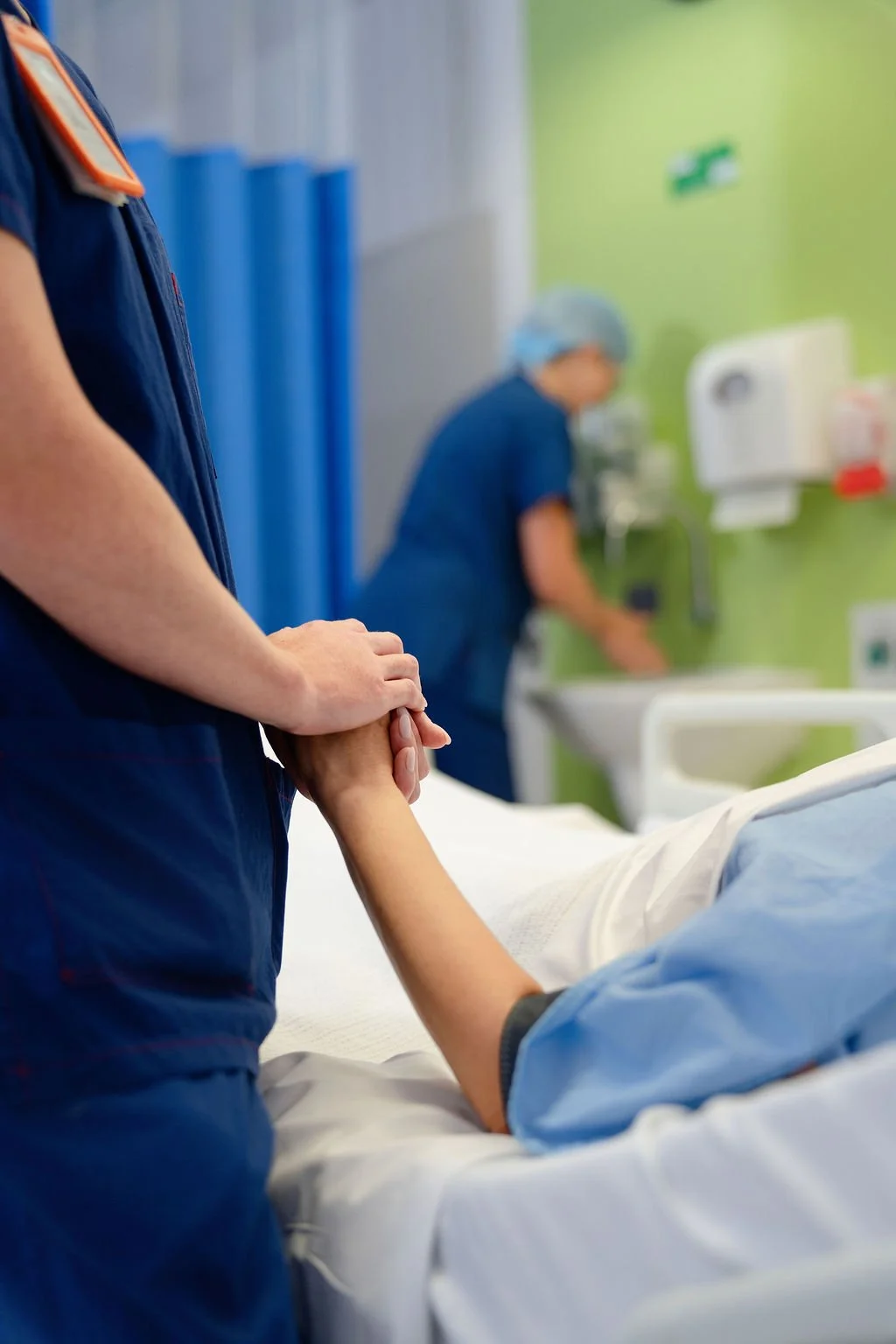 A nurse in navy scrubs gently holds a patient's hand as they wait in a hospital bed, with a second clinician working in the background and blue privacy curtains and green accent walls visible
