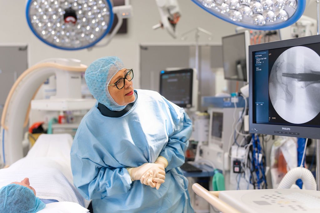 A female orthopaedic surgeon in surgical gown and glasses leans towards a Philips fluoroscopy monitor displaying a spinal X-ray, with theatre equipment and an anaesthetised patient visible in the background.