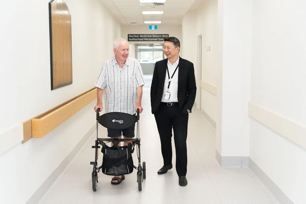 An elderly male patient using a walking frame and a male orthopaedic surgeon in business attire walk side by side along a bright hospital corridor, both relaxed and in conversation
