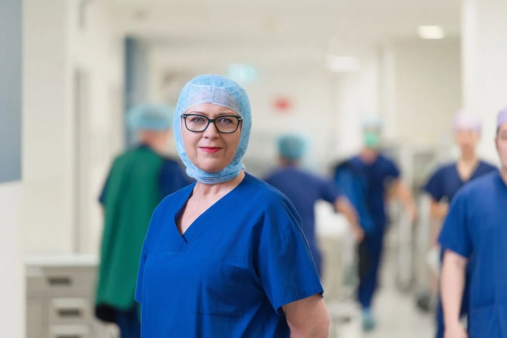 A female nurse in blue scrubs and surgical cap stands facing camera in a hospital corridor, with blurred surgical staff visible behind her in an active clinical environment.