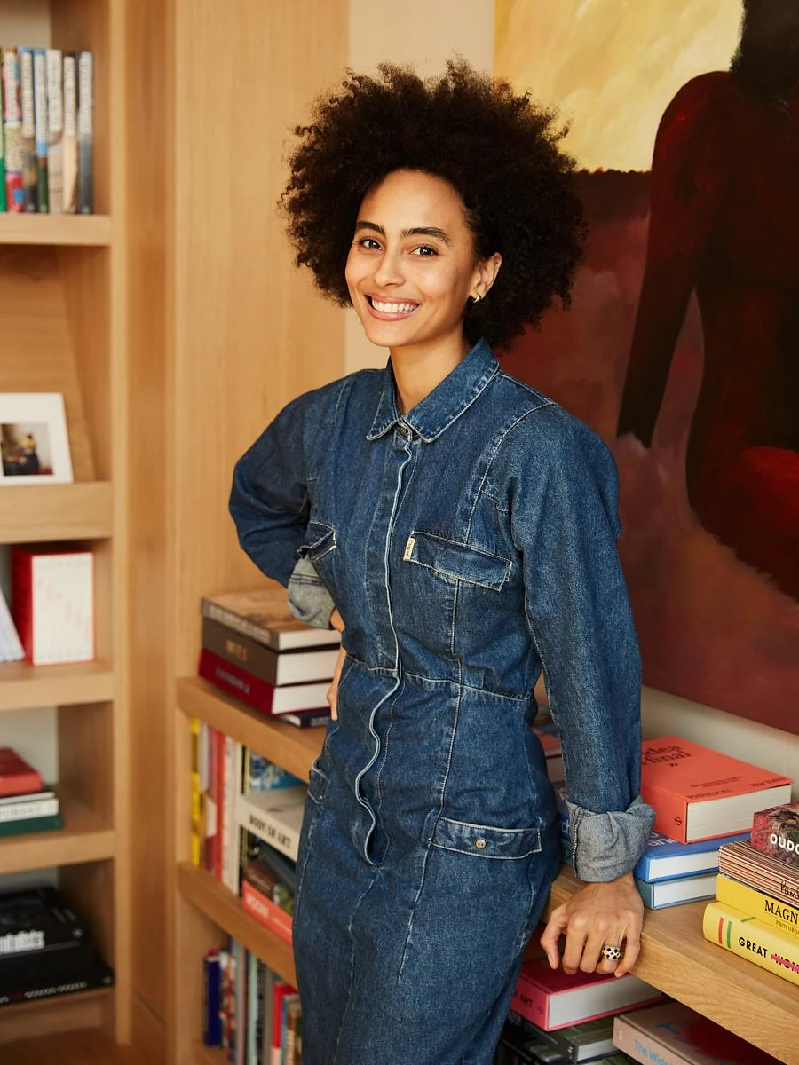 A woman with curly hair smiling in front of bookshelf and artwork, wearing denim dress with rolled-up sleeves.