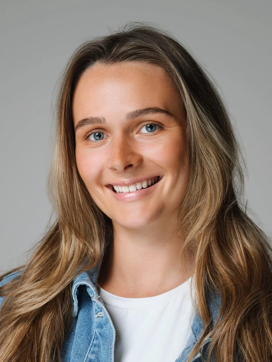 Close-up of a young woman with long brown hair, blue eyes, and a bright smile, wearing a white shirt and denim jacket against a plain gray background by Match Production.