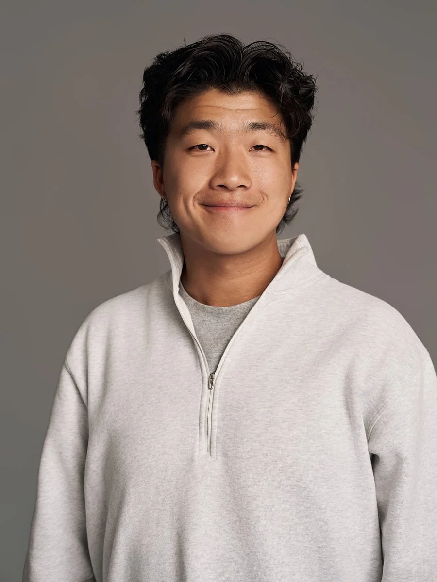 A young Asian man with dark wavy hair and a slight smile, wearing a light gray zip-up sweater against a plain gray background by Match Production.