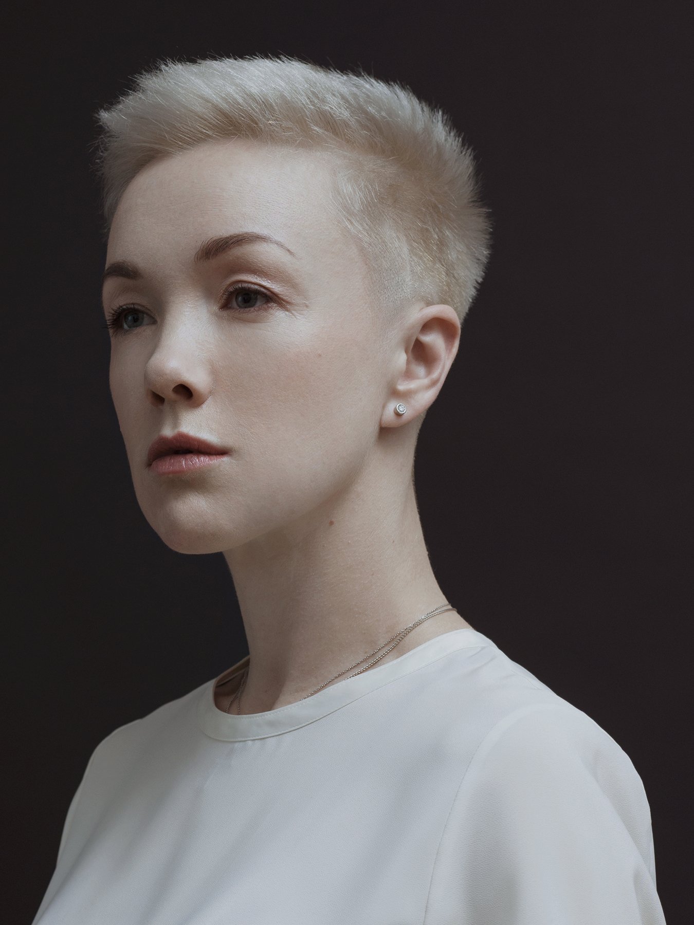 Person with short blonde hair and white top on dark background