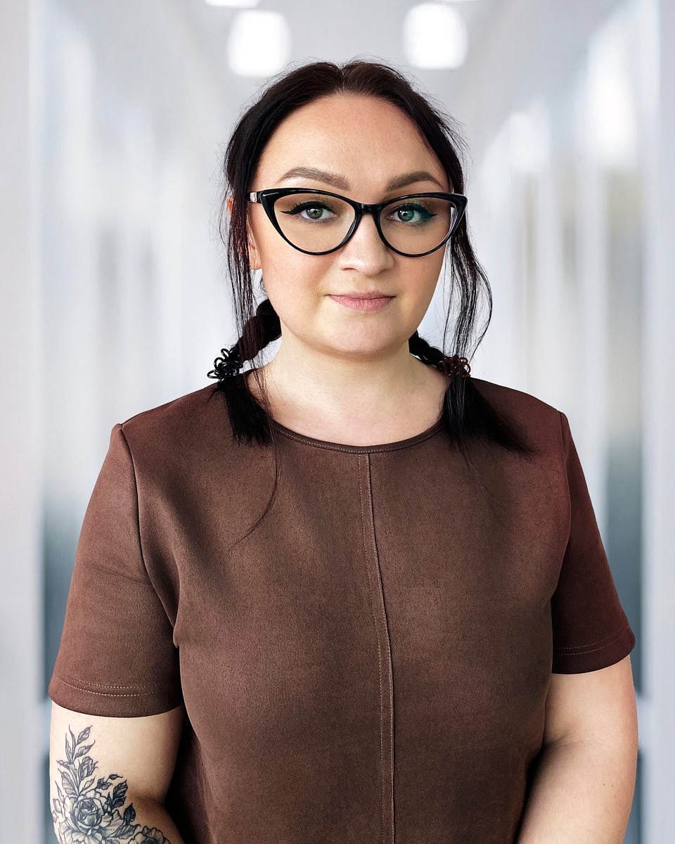 A woman with dark hair in braids, wearing glasses and a brown shirt, standing in a hallway.