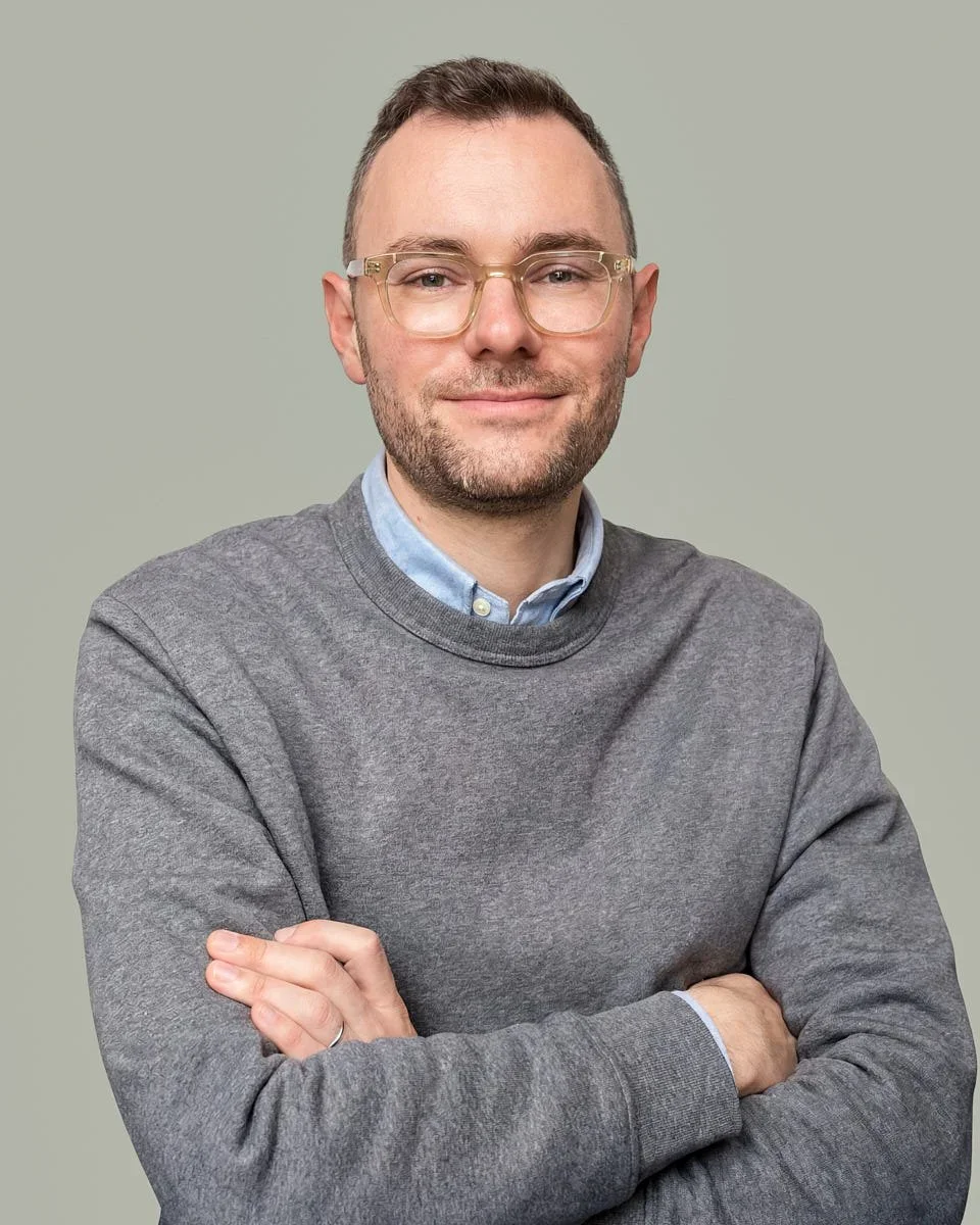 A man with glasses, a beard, and short hair, wearing a gray sweatshirt over a light blue collared shirt, standing against a plain light-colored background with his arms crossed and a slight smile.