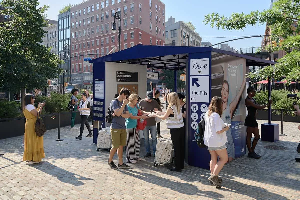 People at a New York City subway station entrance called The Pits, with a large advertisement for Dove on the side, some waiting in line and others taking photos, surrounded by trees and buildings.