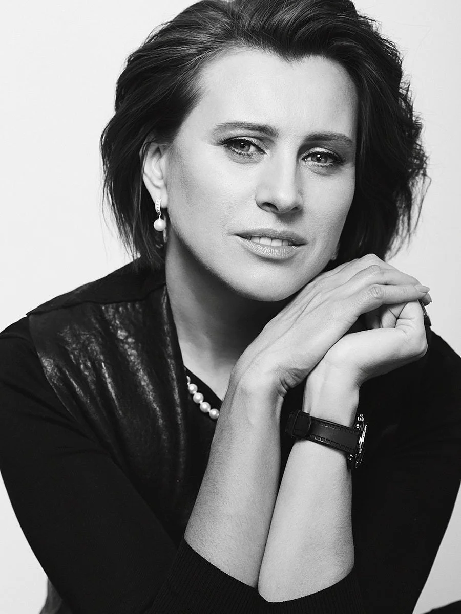 Black and white portrait of a woman with short dark hair, wearing pearl earrings, a pearl necklace, a black leather jacket, and a watch, with her hands gently resting on her chin.
