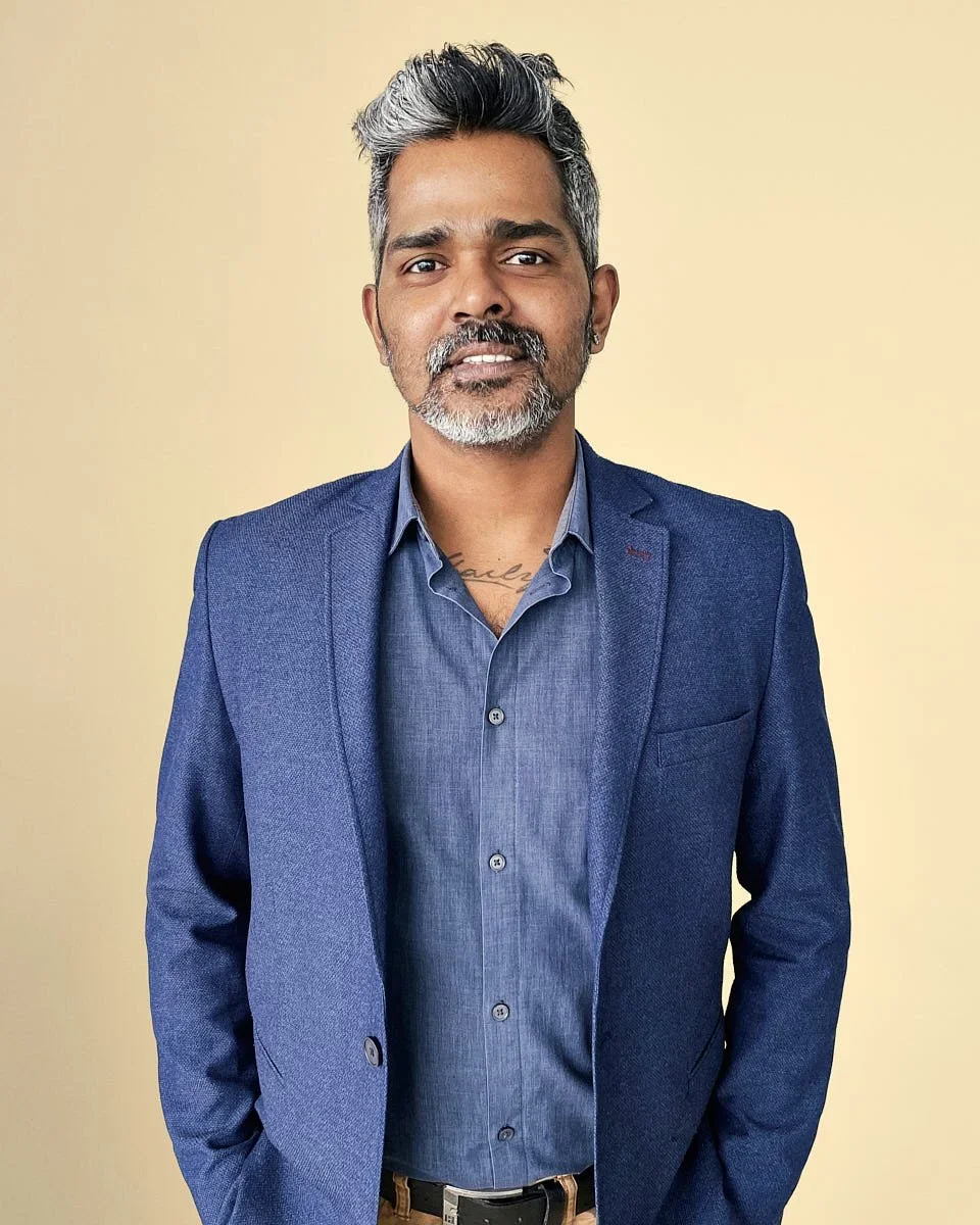A man with salt-and-pepper hair and beard wearing a blue suit and shirt, standing against a yellow background.