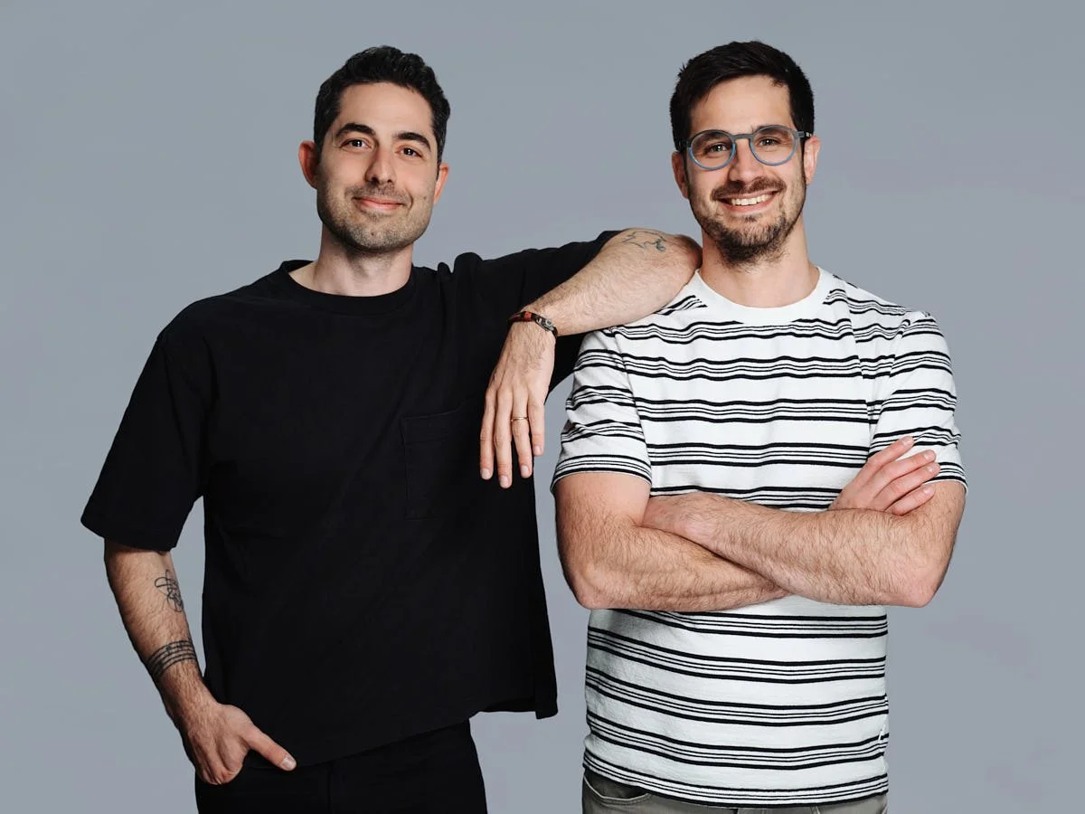 Two smiling young men with dark hair and glasses, one with a beard, standing against a gray background. One wears a black t-shirt, and the other wears a white and black striped t-shirt. The man in the black t-shirt has his arm resting on the shoulder