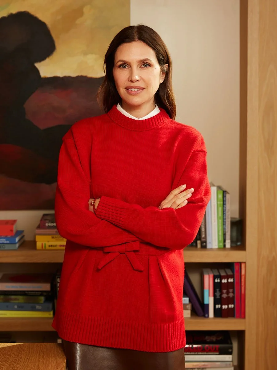 A woman stands in a room with her arms crossed, wearing a red sweater and brown pants, with a bookshelf and a painting in the background.