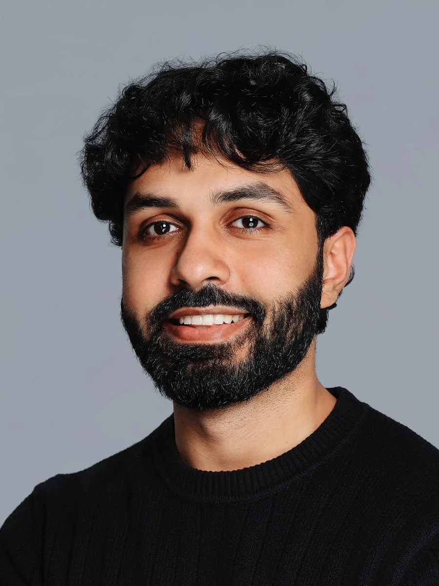 Portrait of a man with dark, curly hair and a full beard, smiling at the camera, wearing a black sweater against a plain gray background by Match Production NYC.