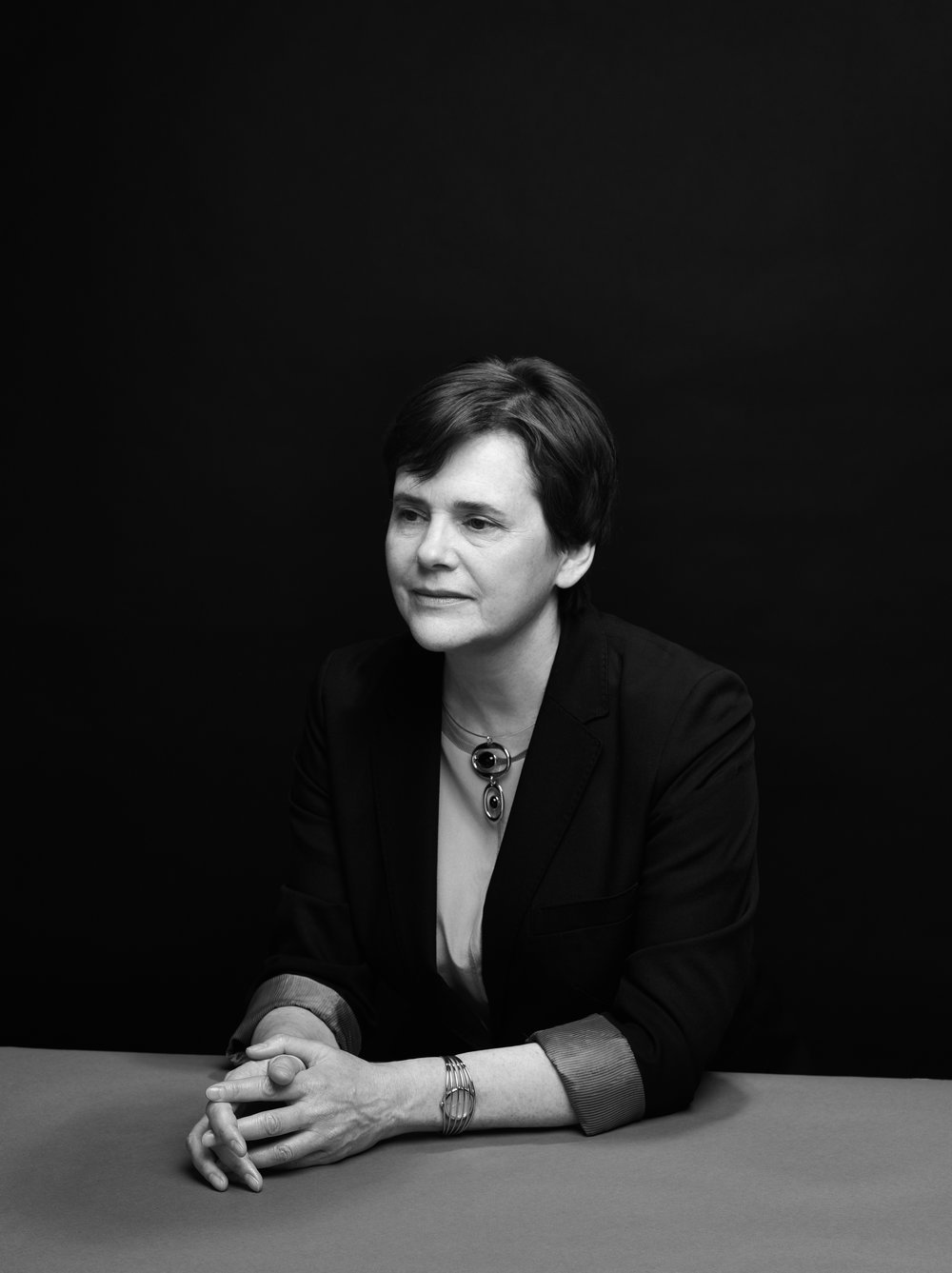 Black and white portrait of a person sitting at a table with hands clasped, wearing a blazer and jewelry, against a dark background.