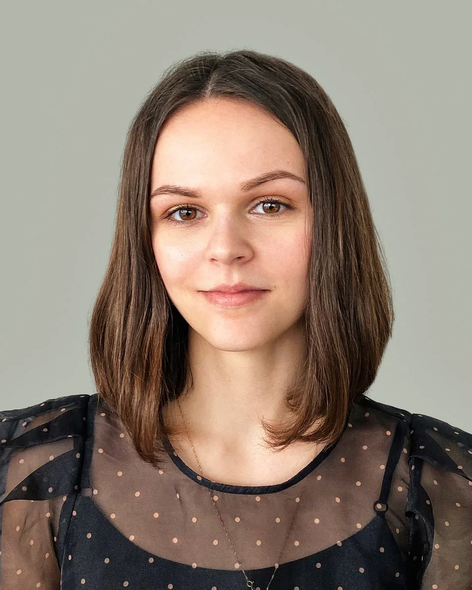 A young woman with brown hair styled in loose waves, wearing a black sheer top with pink polka dots and a delicate necklace, standing against a plain mint green background.