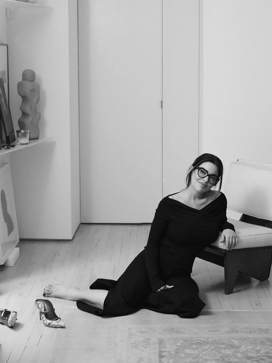 A woman in a black dress and glasses is kneeling on the floor next to a couch in a room with white walls and a wooden floor. She is smiling at the camera.