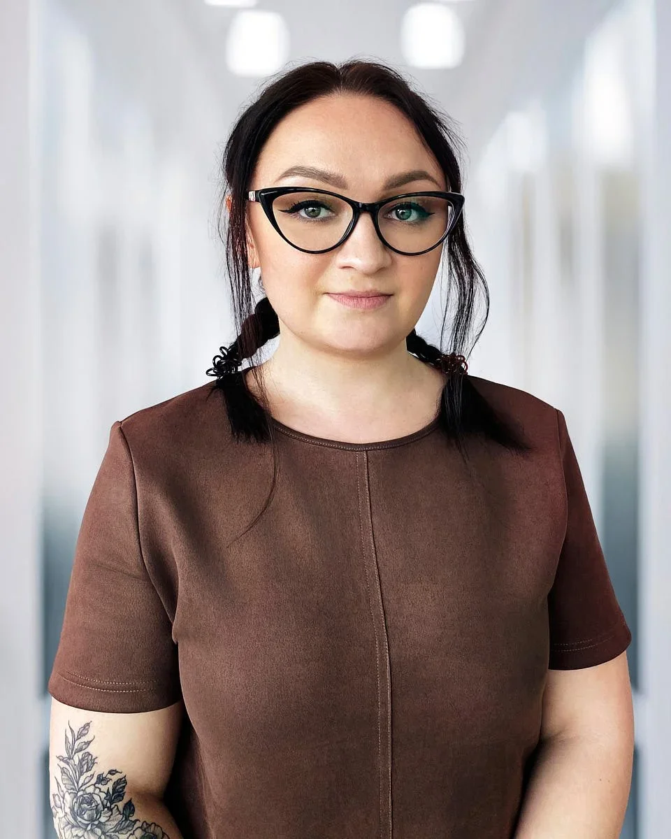 A young woman with dark hair tied into two braids, wearing glasses, a brown shirt, and featuring a floral tattoo on her left arm, standing in a blurred indoor setting.