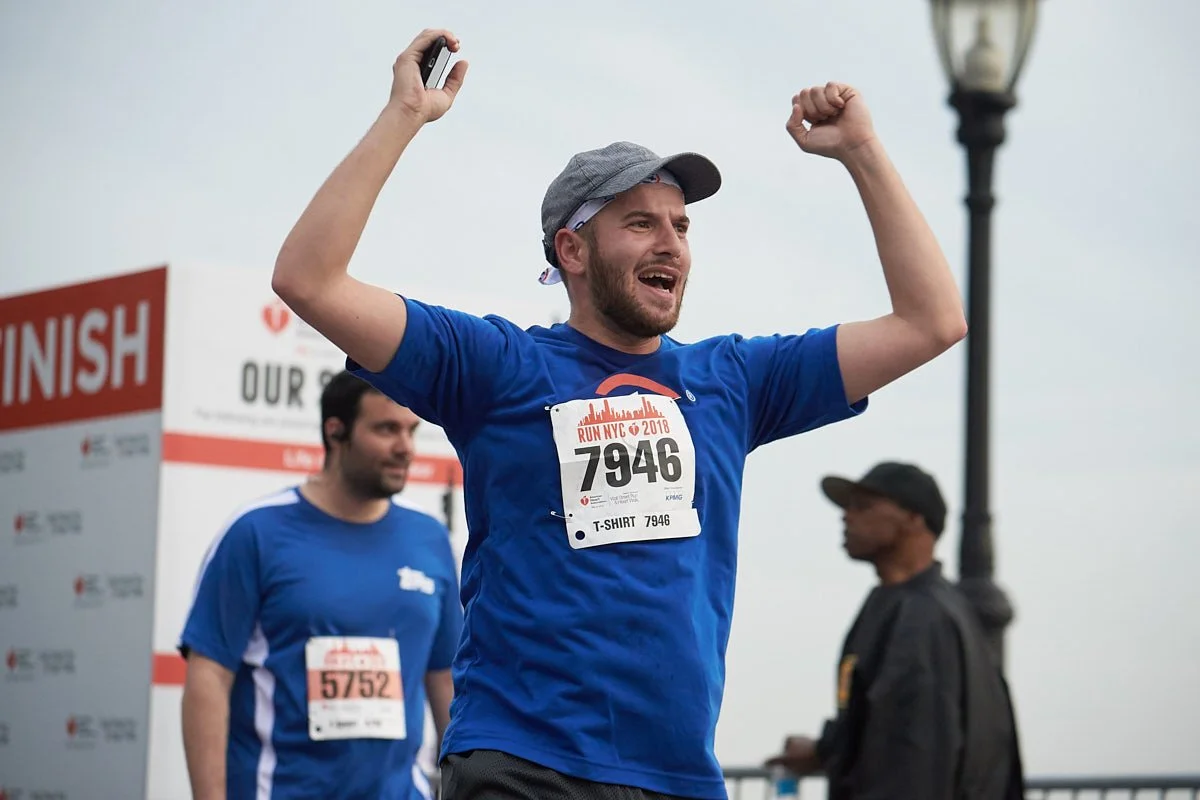 A man wearing a gray cap and blue t-shirt celebrating after finishing a race, with his right arm raised in victory, at a marathon event with a finish line banner in the background.