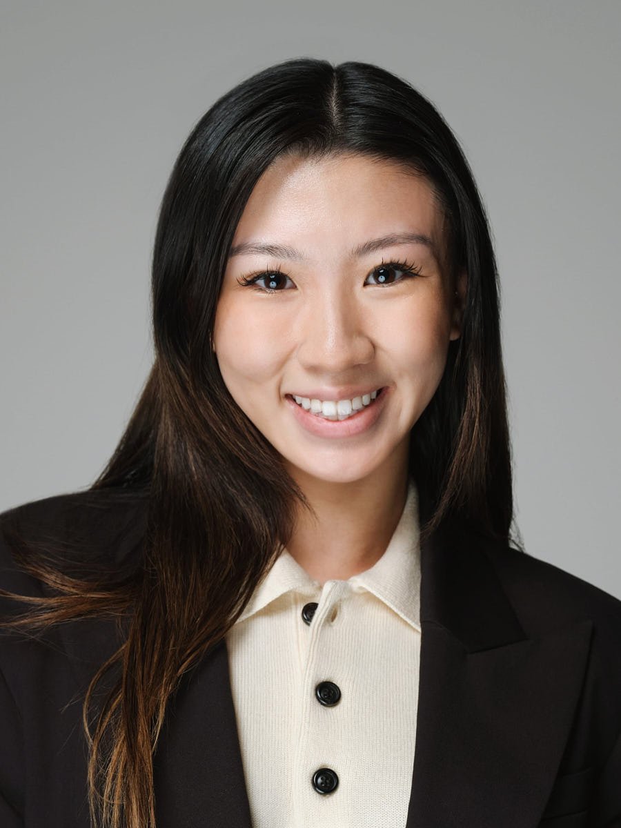 A young woman with long black hair, smiling, wearing a beige shirt and black blazer against a gray background by Match Production.